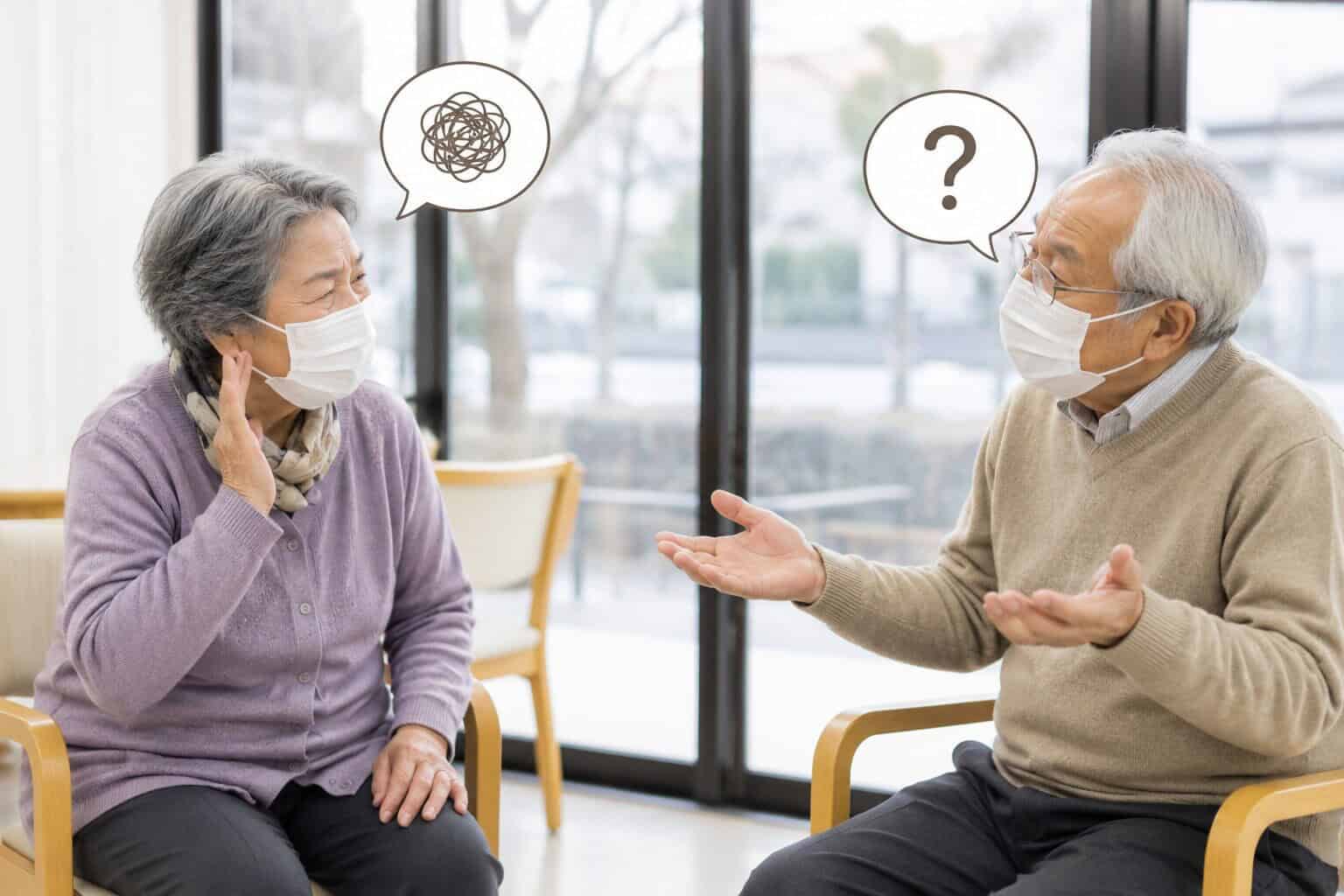 「マスクを着けた高齢者同士が距離をとって会話しているが、お互い声が届きにくく困っている様子を描いたイラスト。冬の感染対策下での“聞こえにくさ”を表現。」